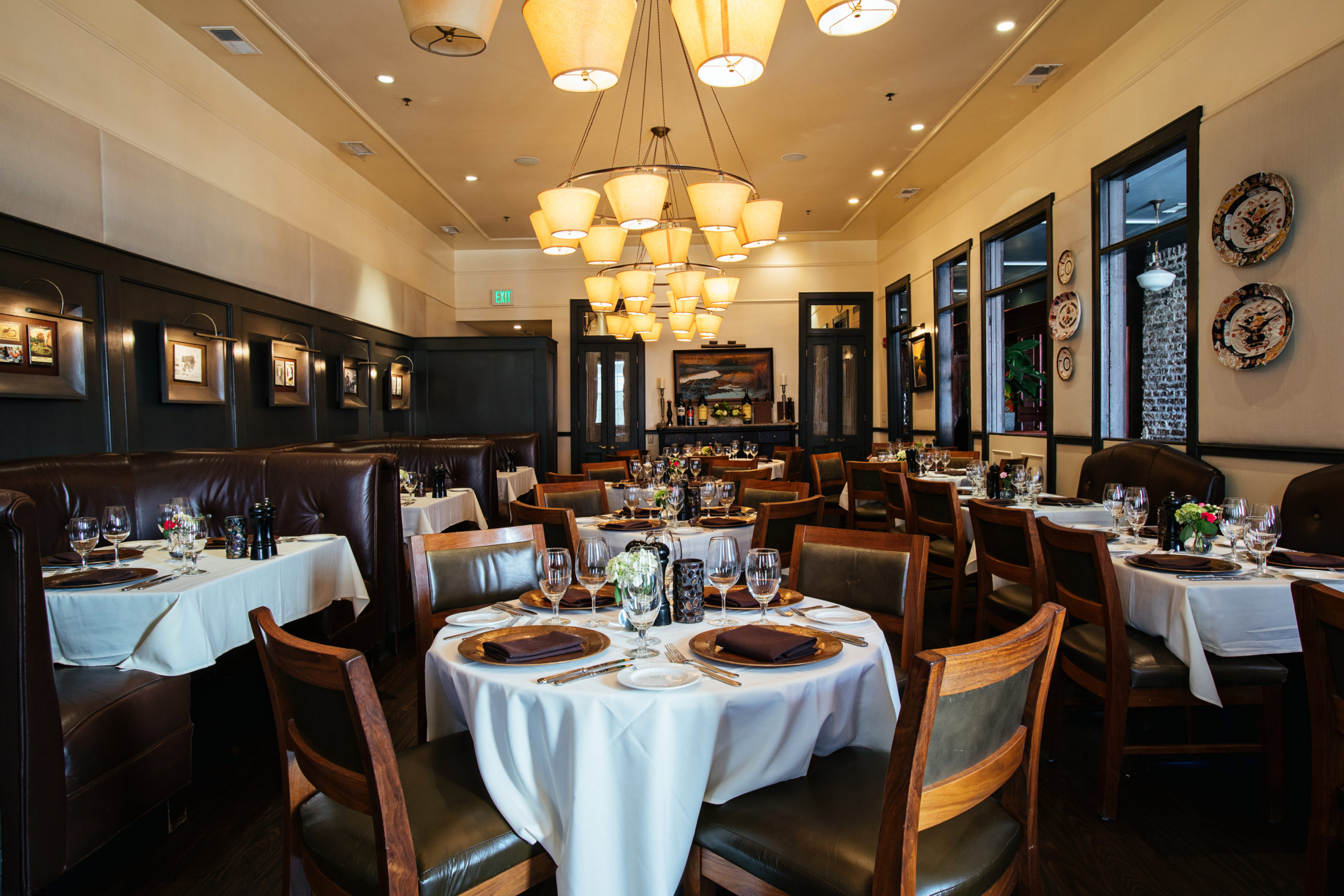 Large restaurant dining room with warm lighting from large overhead chandeliers, leather and wood chairs, table linens and nice place settings