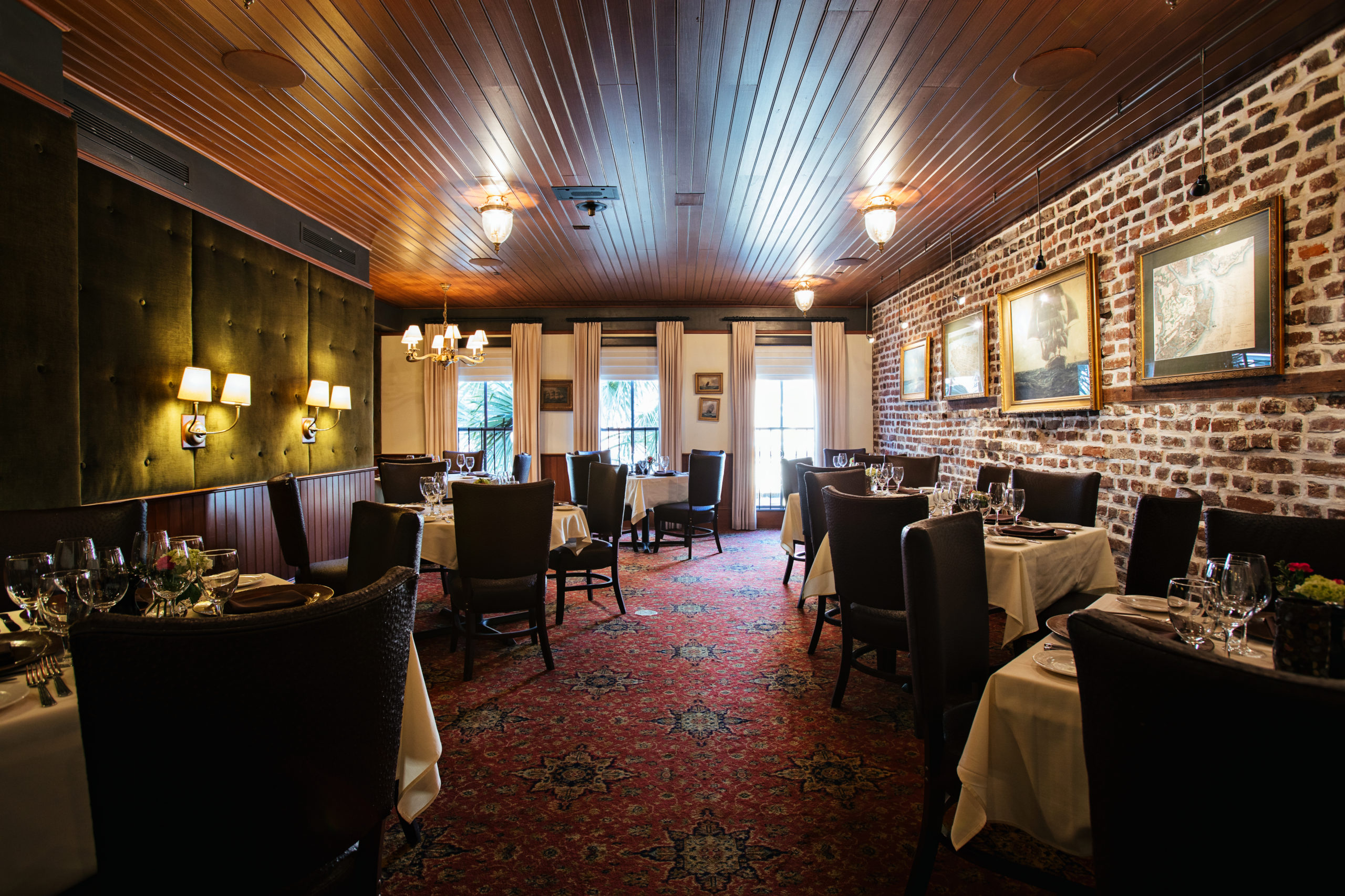 Large restuarant dining room with high back dining hairs, patterned red rug, exposed brick wall, and a velvet green feature wall with shaded sconces