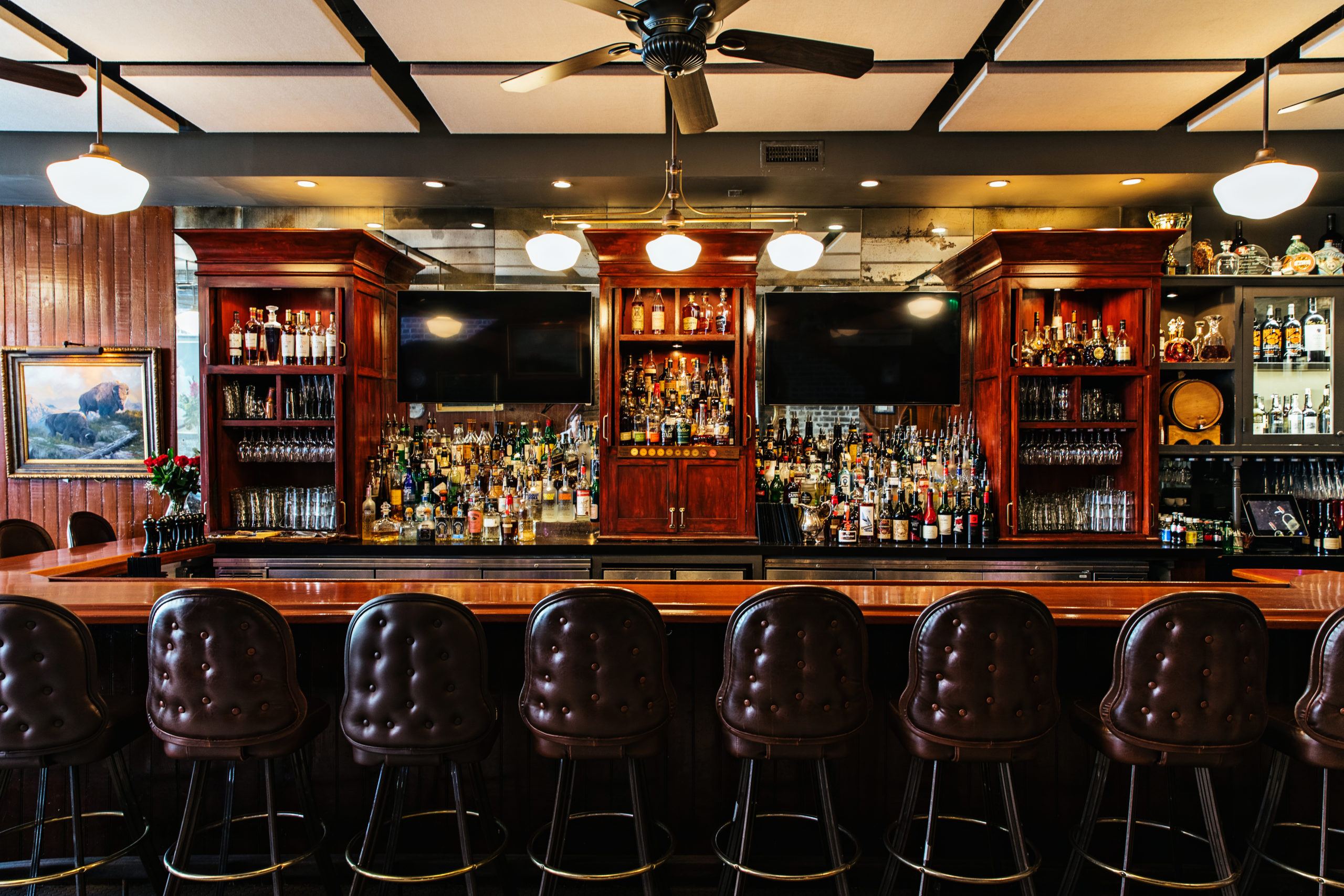 Fully stocked bar made of dark wood, leather tufted chairs and frosted pendant lights